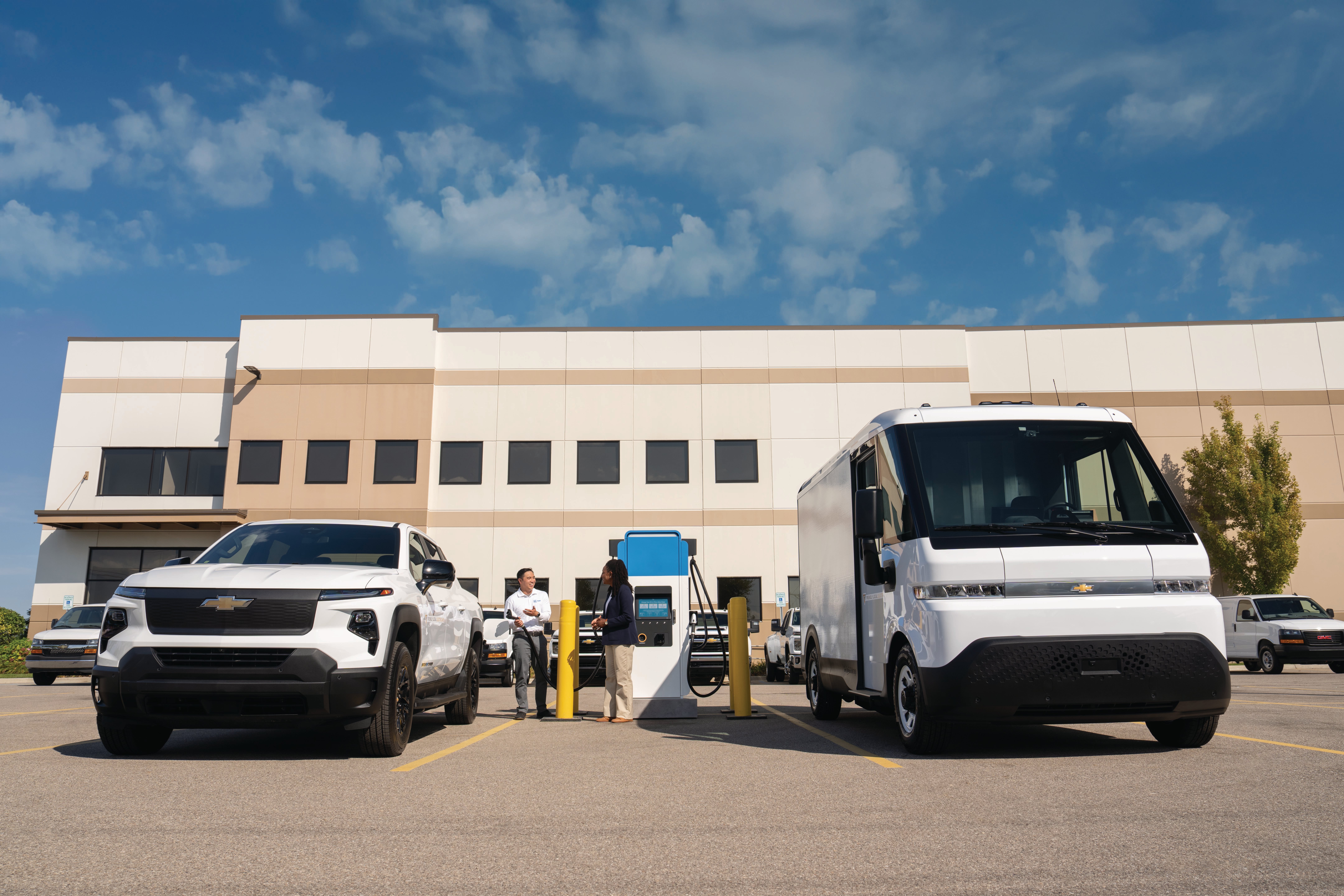 White Chevrolet Silverado EV truck and BrightDrop van parked at a commercial EV charging station.
