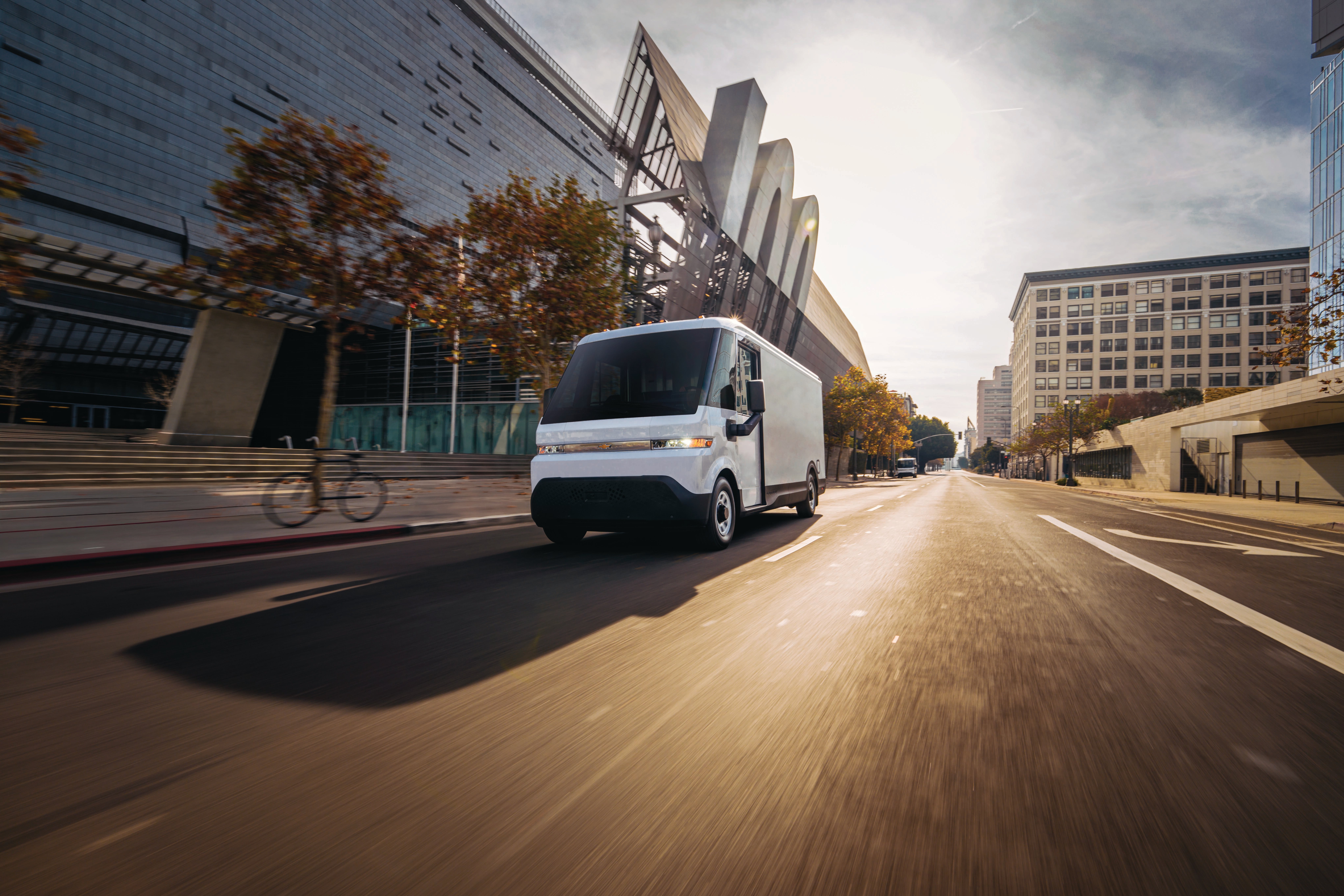 White modern electric delivery van driving down a sunlit city street with modern architecture in the background.
