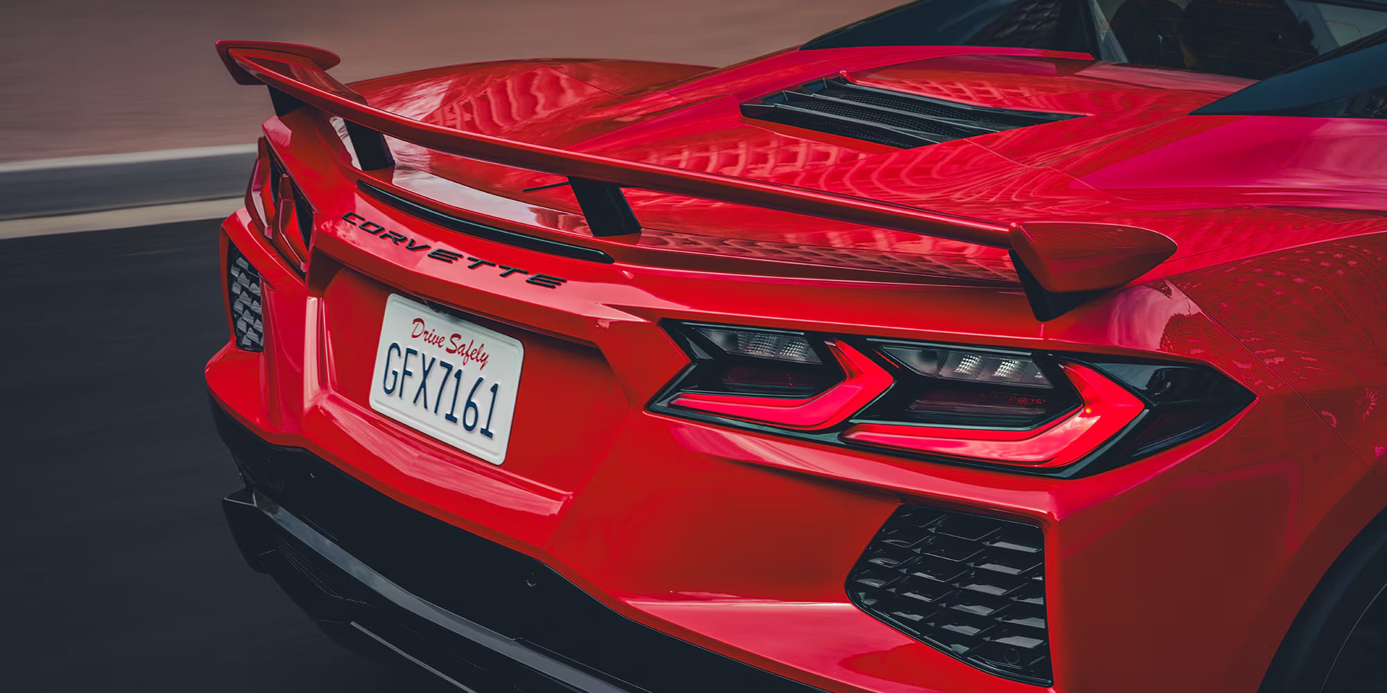 Rear view of a red Chevrolet Corvette C8 driving on a road showing taillights, spoiler, and license plate.