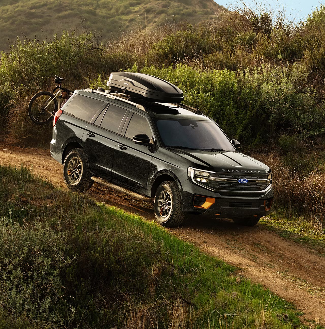 A dark green Ford Expedition with a roof cargo carrier and rear-mounted bike drives on a dirt hill trail.