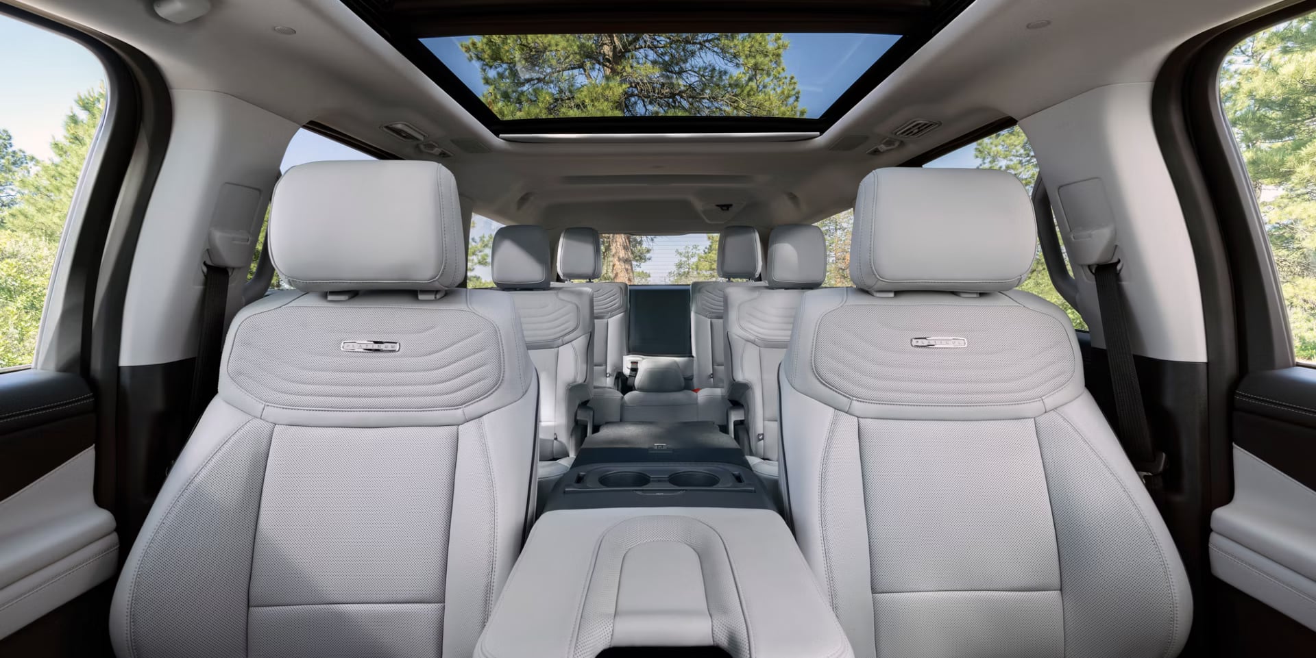Interior view of a luxury vehicle featuring light gray leather seating and a large panoramic sunroof looking out at trees.