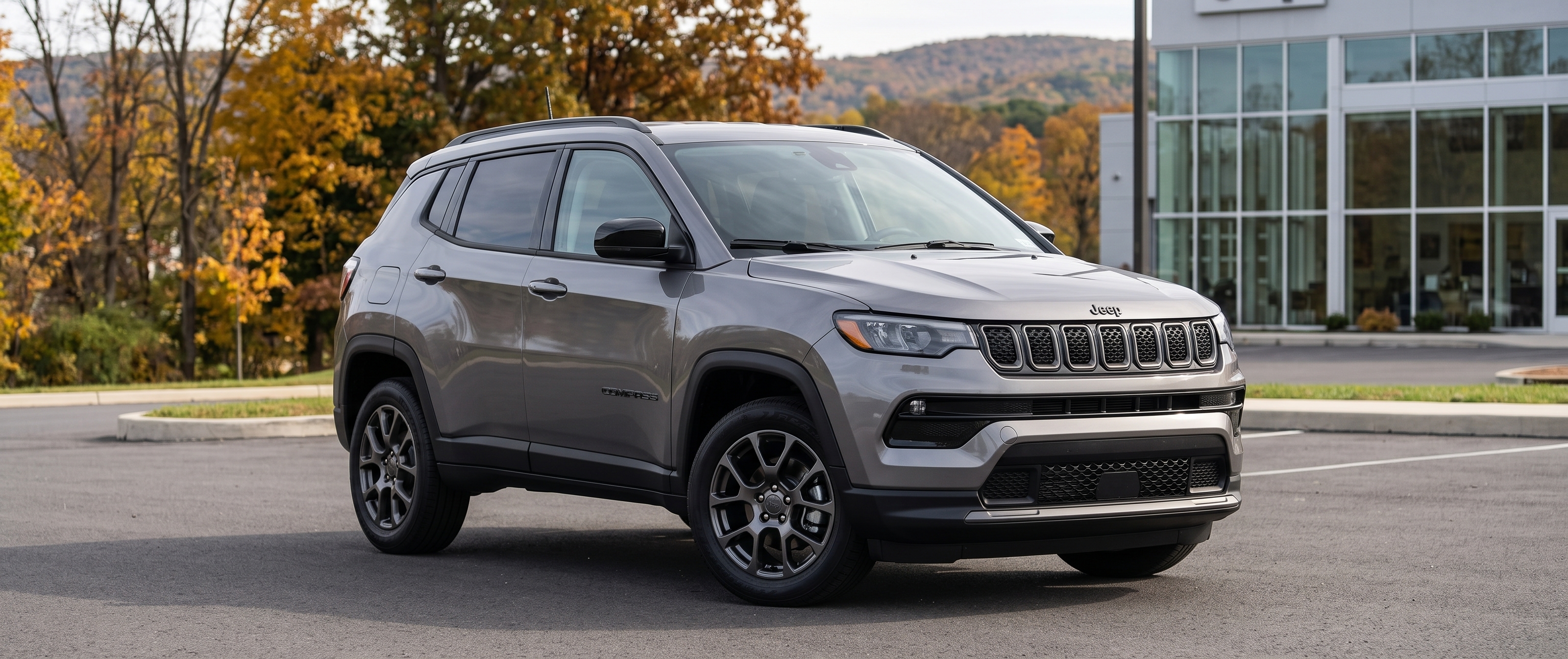 Grey Jeep Compass SUV parked in a car dealership lot with autumn trees in the background.