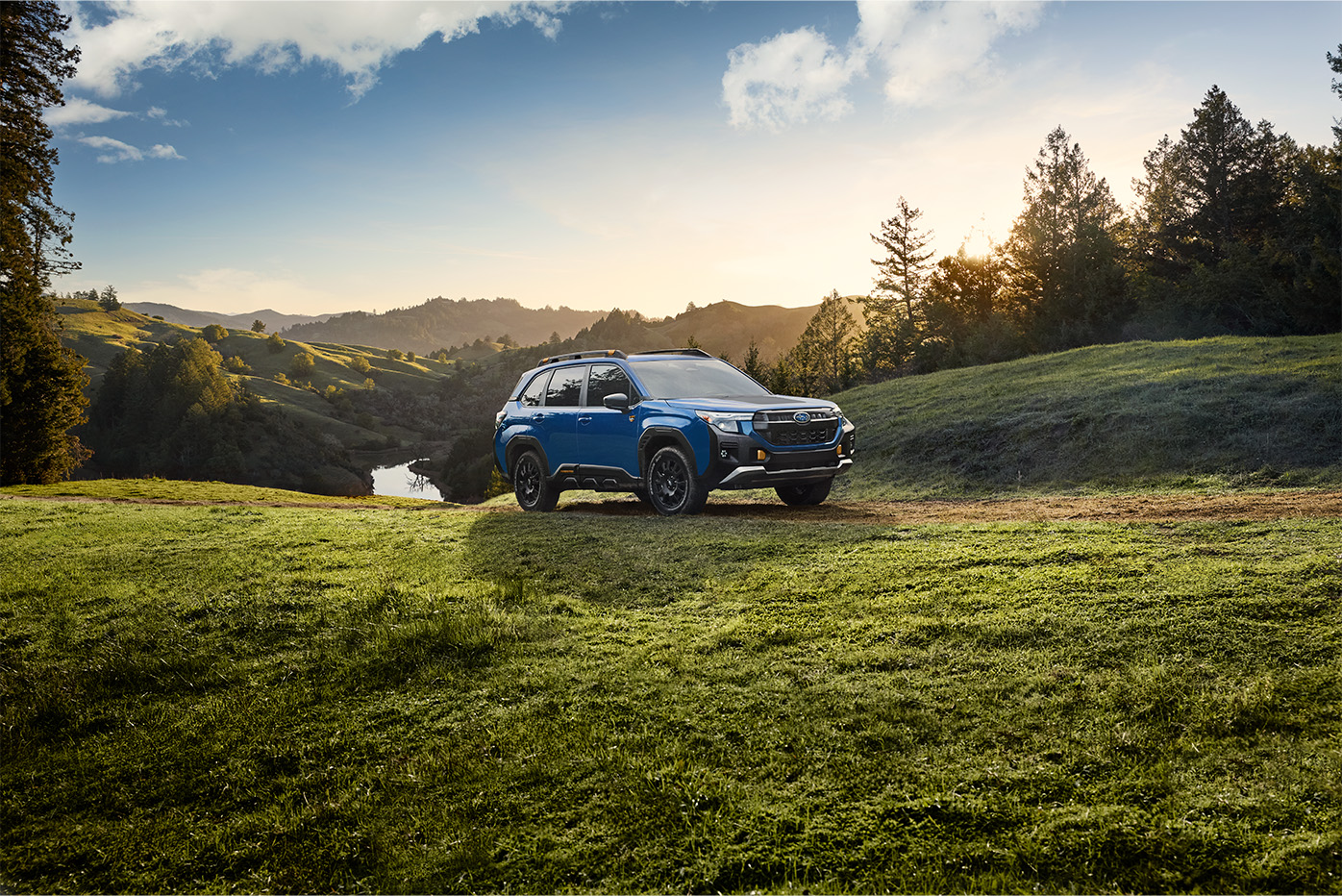 Blue Subaru SUV parked on a dirt trail on a grassy hill during sunset with scenic mountain views.