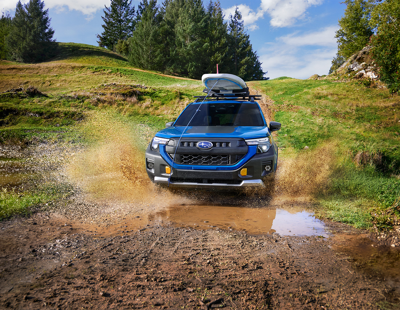A blue Subaru SUV driving through a muddy puddle on an off-road path with a surfboard on the roof rack.