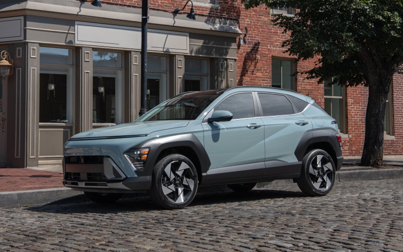 Light sage green Hyundai Kona SUV parked on a cobblestone street beside a brick building.