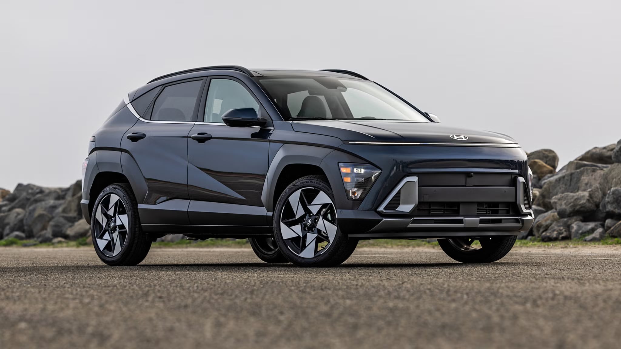 Front three-quarter view of a dark blue Hyundai Kona SUV parked on an asphalt road near rocks.