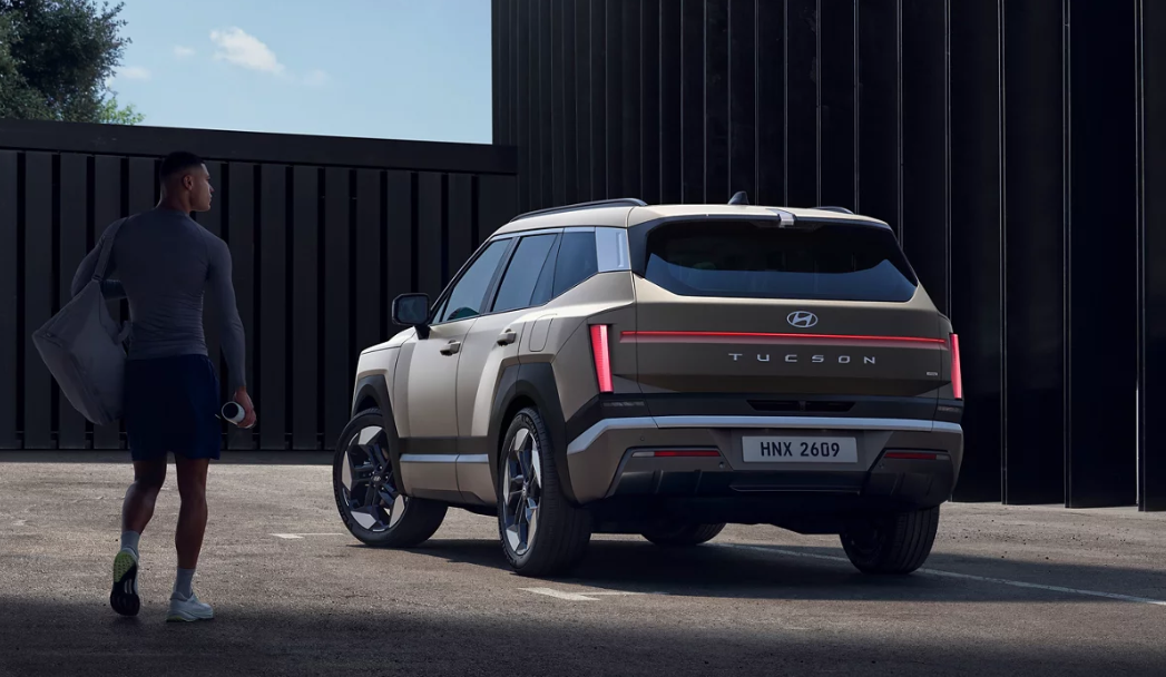 Rear view of a beige Hyundai Tucson SUV parked near a modern building as a man carrying a gym bag walks past.