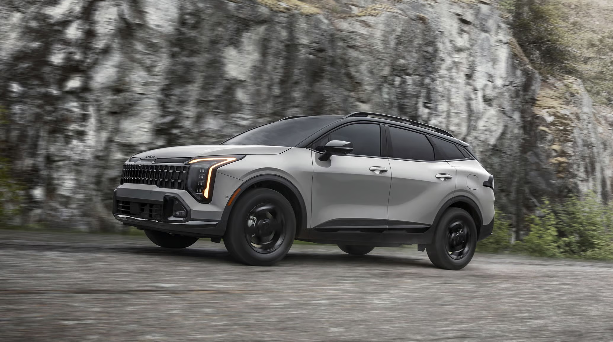 Silver Kia Sportage driving along a paved road beside a rocky cliff.