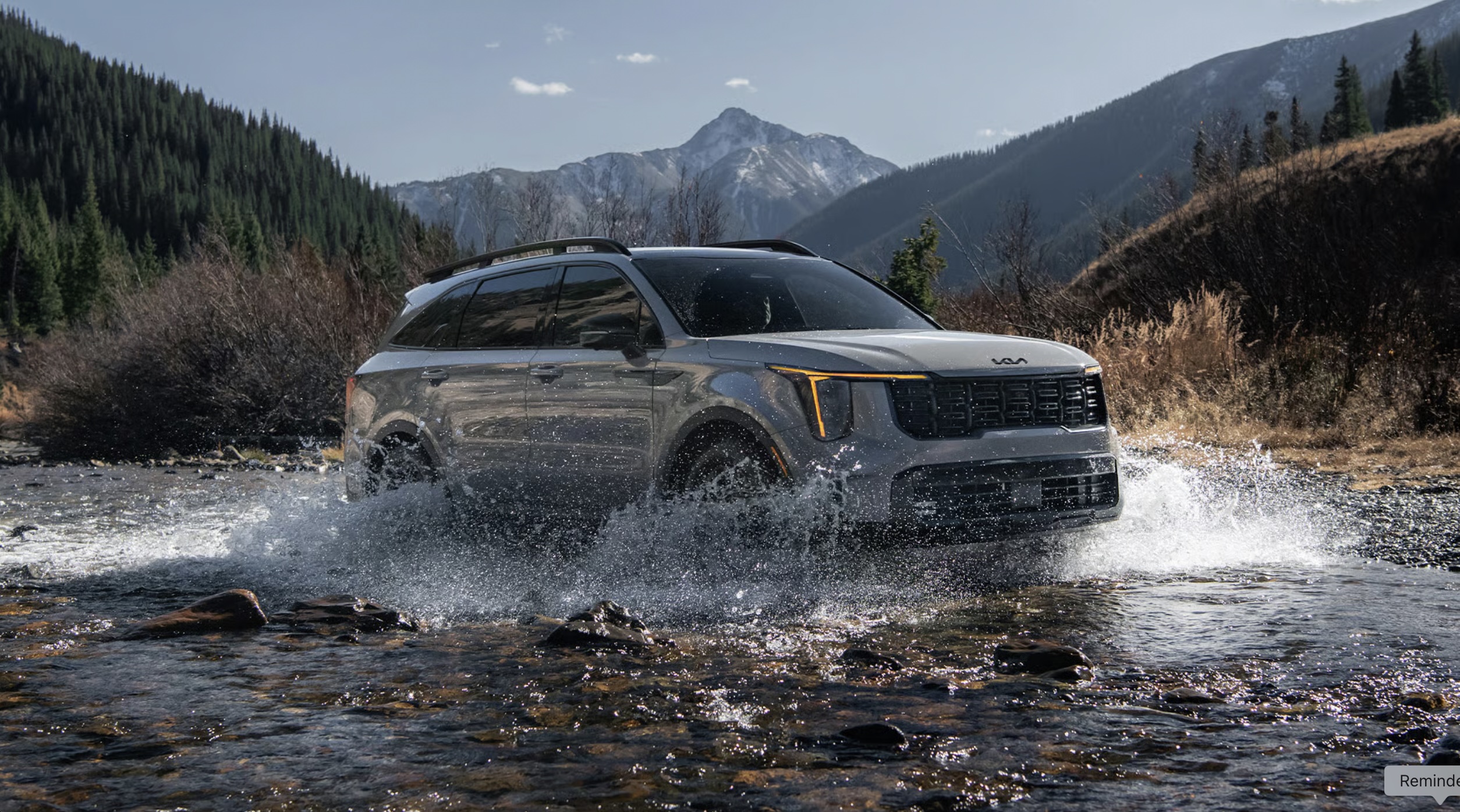 Silver Kia Sorento driving through a shallow rocky stream with mountains in the background.