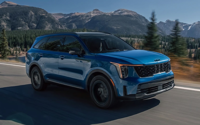 Blue Kia Sorento driving on a scenic highway surrounded by mountains and pine trees.