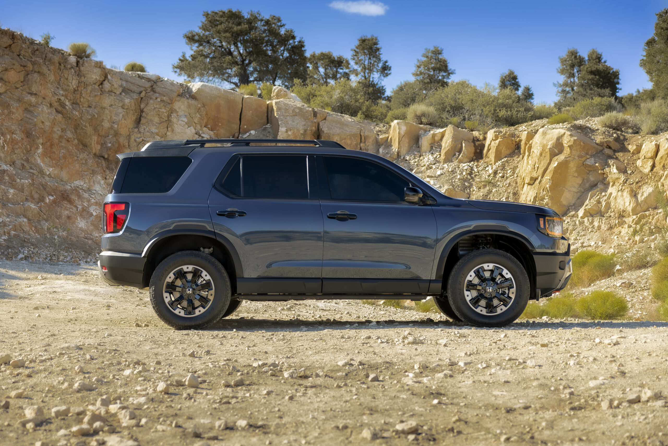 Side profile of a dark blue Honda SUV parked on a rocky off-road terrain with cliff background.