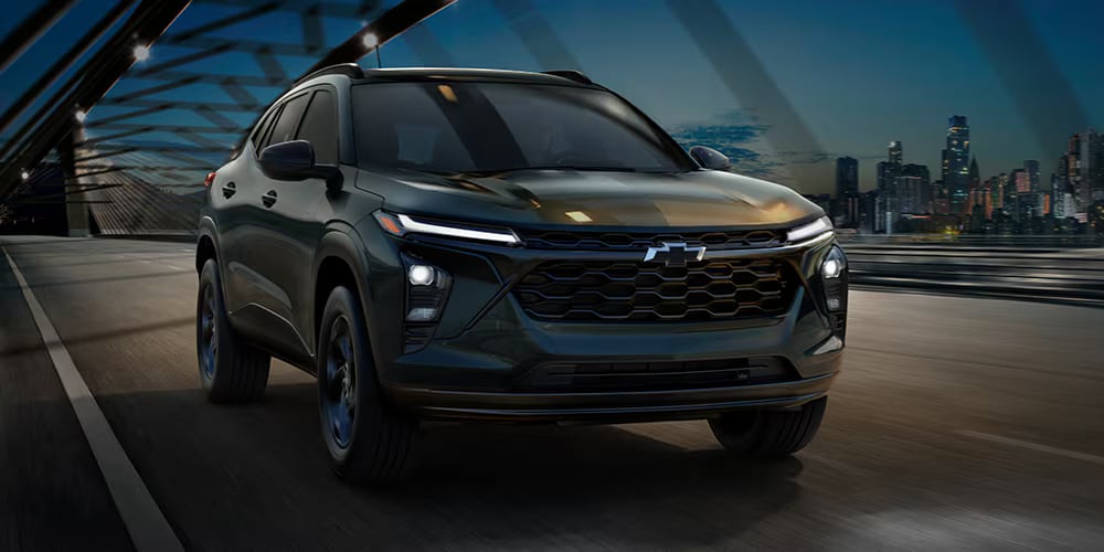 Front view of a dark green Chevrolet Trax driving on a bridge at dusk with a city skyline behind it.