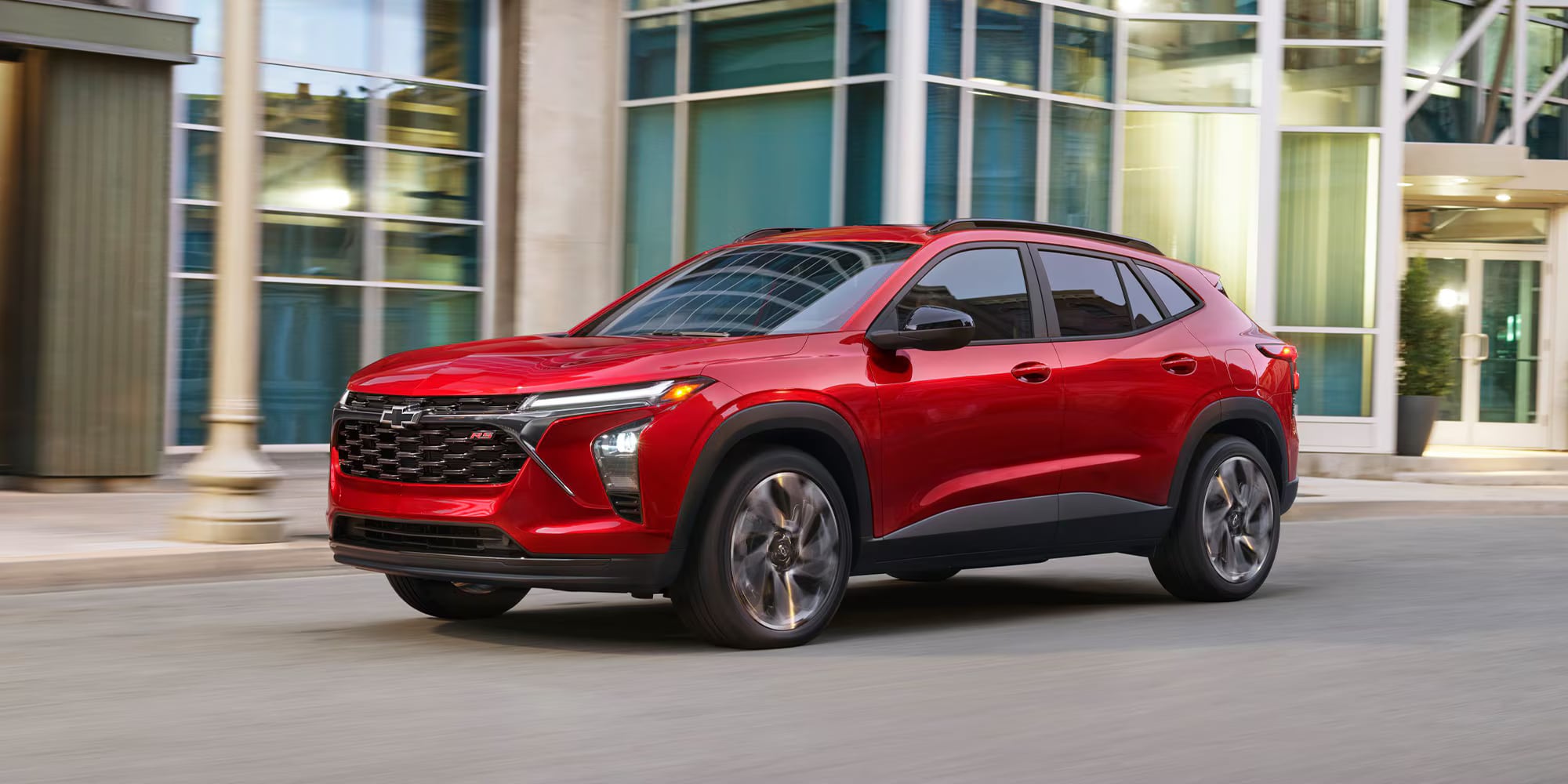 Red Chevrolet Trax RS crossover driving past modern glass buildings on a city street.