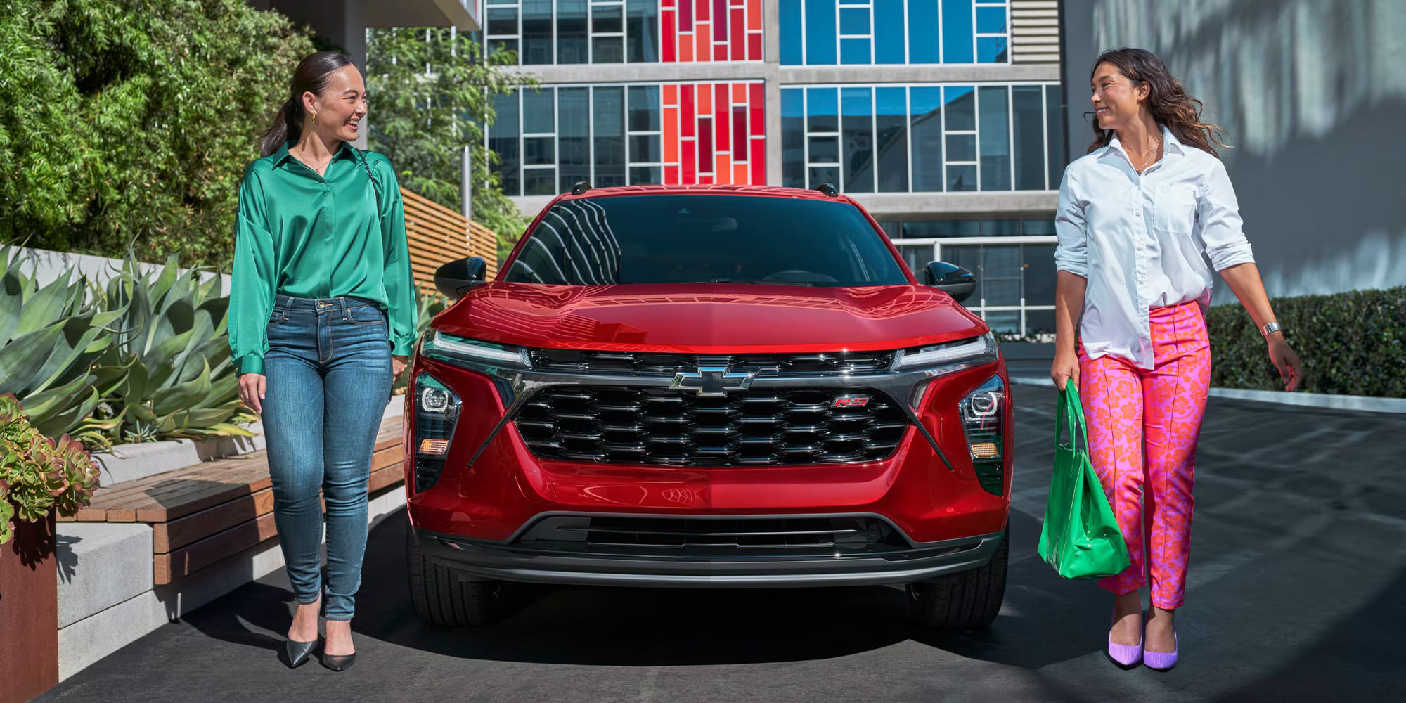Two women standing on either side of a bright red Chevrolet Trax RS SUV outside a modern building.