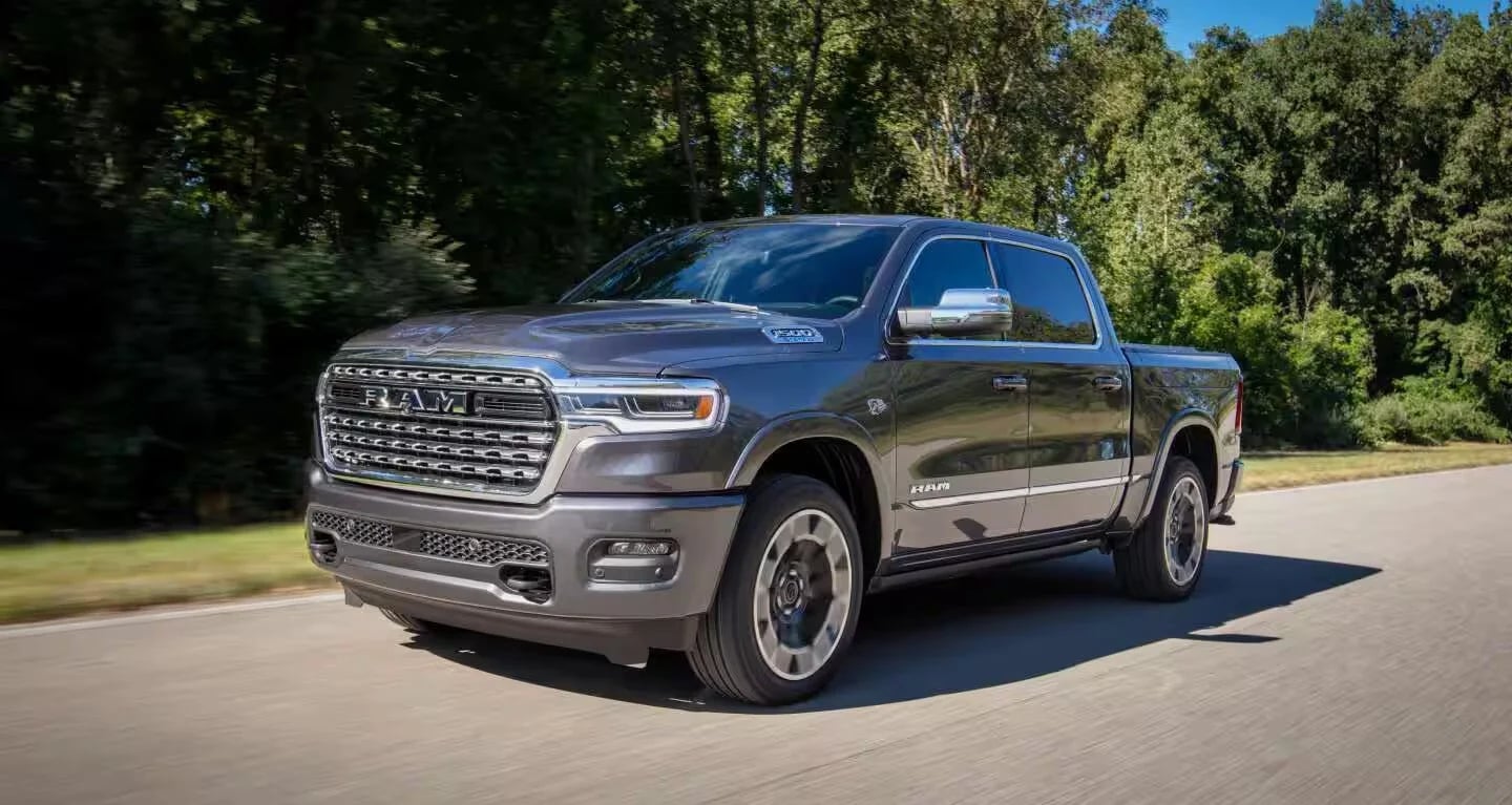 Dark grey Ram 1500 pickup truck driving on an asphalt road next to lush green trees.