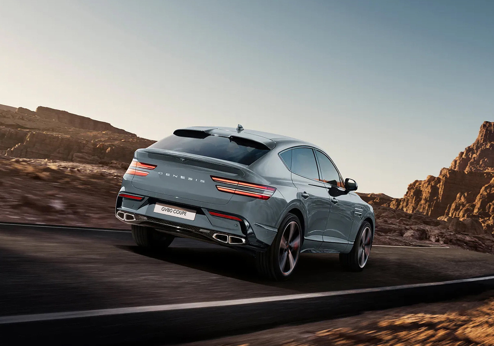 Rear three-quarter view of a gray Genesis GV80 Coupe driving on a scenic road