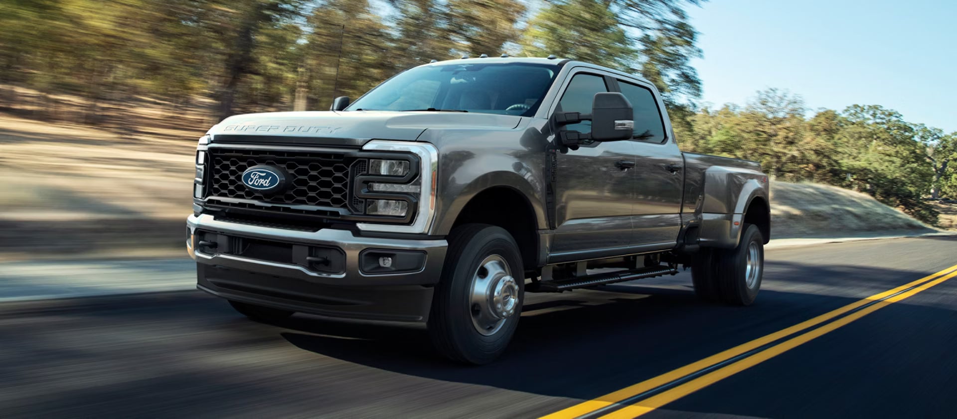 A metallic gray Ford Super Duty dually pickup truck driving along a paved road with trees in the background.