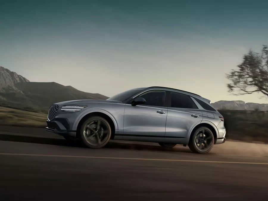 Silver Genesis GV70 luxury SUV driving dynamically along a scenic mountain road at dusk.