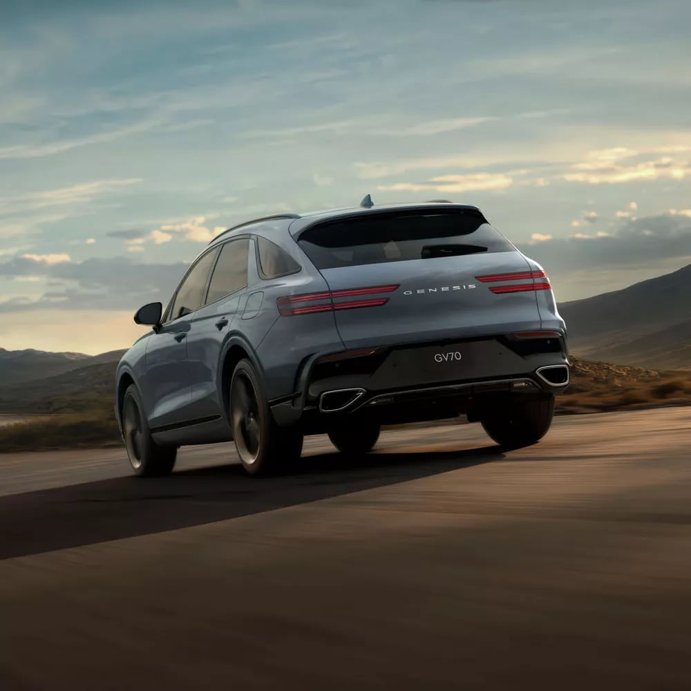 Light blue Genesis GV70 SUV driving along an open highway with mountains under a cloudy sunset sky.