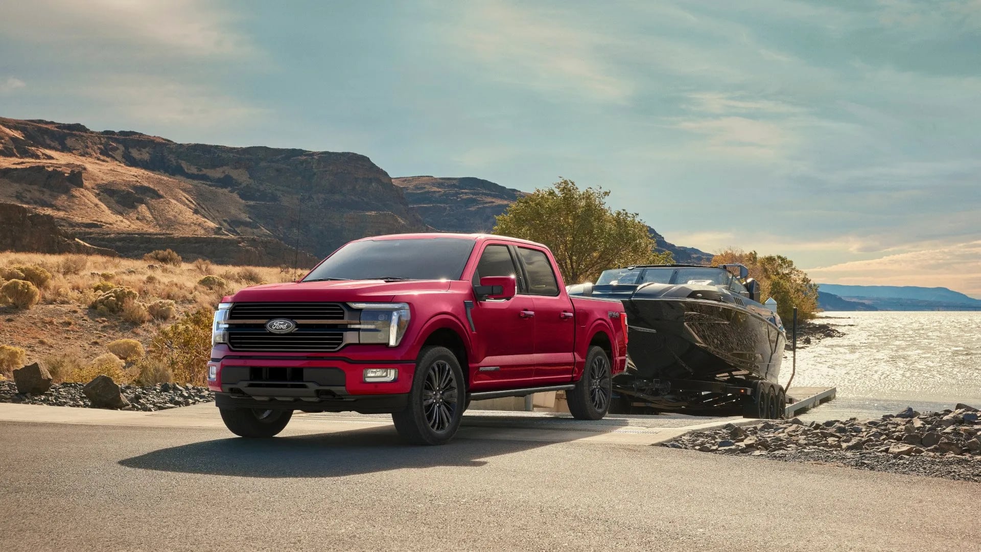 Red Ford F-150 pickup truck towing a black powerboat on a trailer at a scenic lake boat ramp.