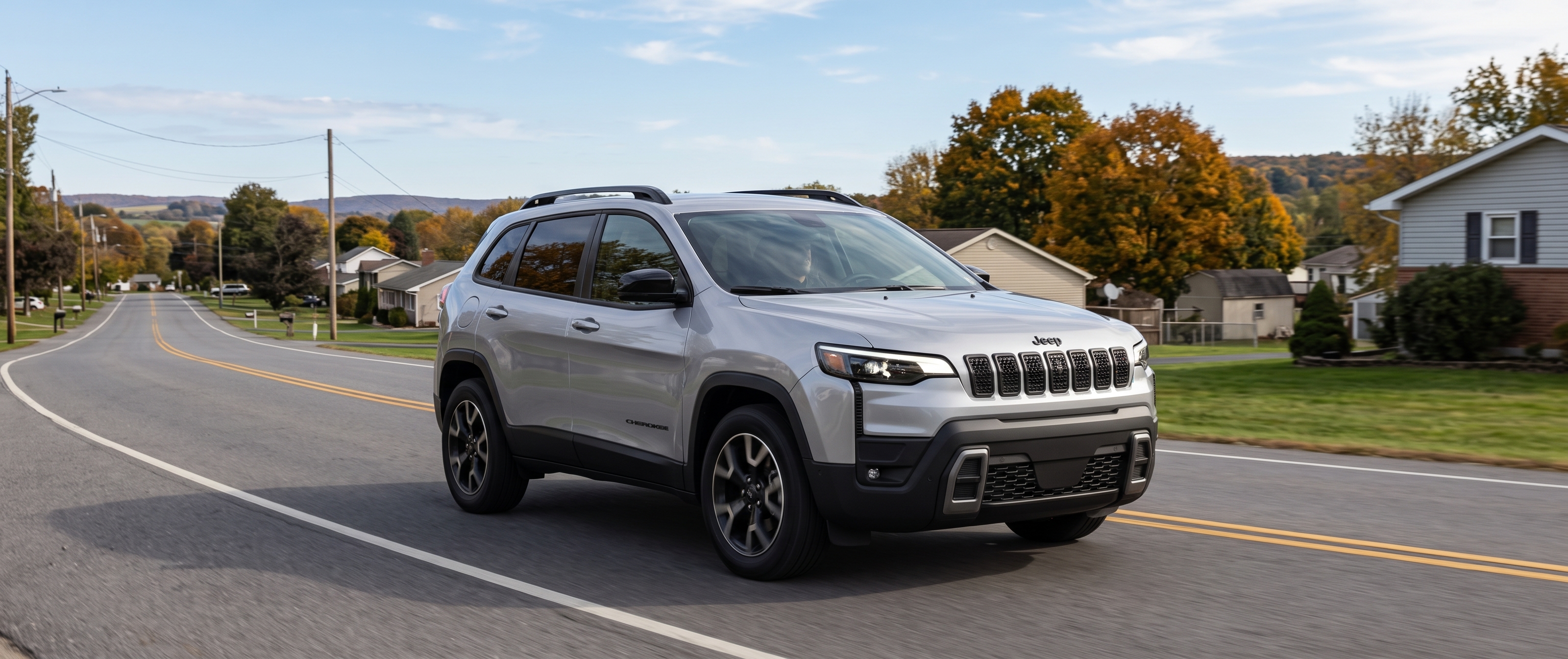 Silver Jeep Cherokee driving along a suburban road lined with autumn trees and houses.