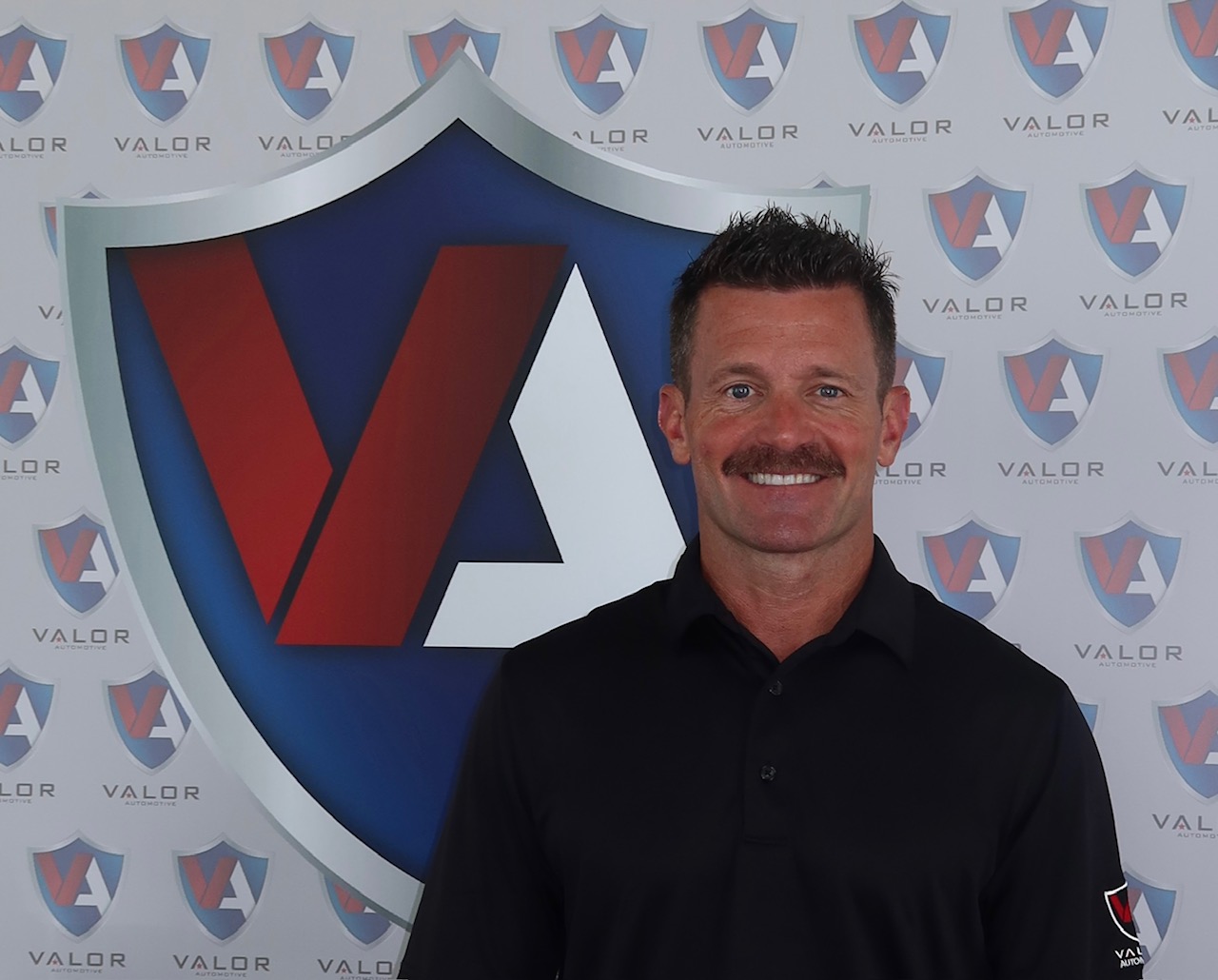 Smiling man with a mustache wearing a black polo shirt in front of a Valor Automotive shield backdrop.
