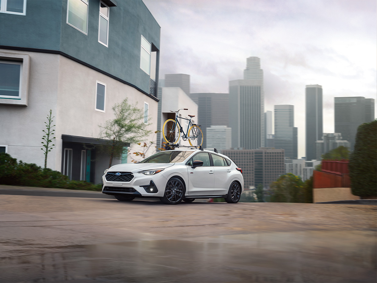 A white Subaru hatchback with a bicycle mounted on its roof rack parked on a hill in front of a city skyline.