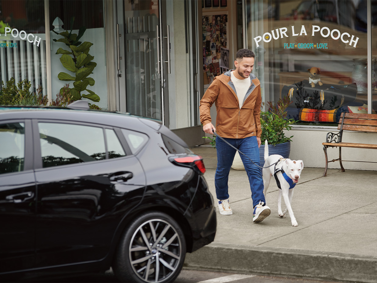 A smiling man walks a white dog on a leash on a sidewalk past a black car in front of a pet store.