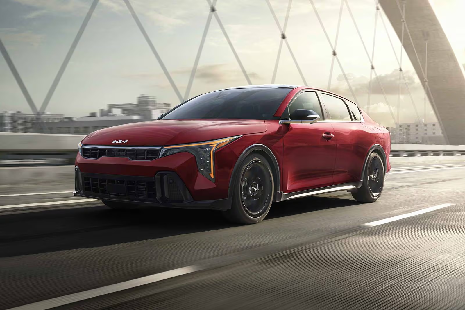 Red 2026 Kia K4 sedan driving across a suspension bridge with a city skyline in the background.