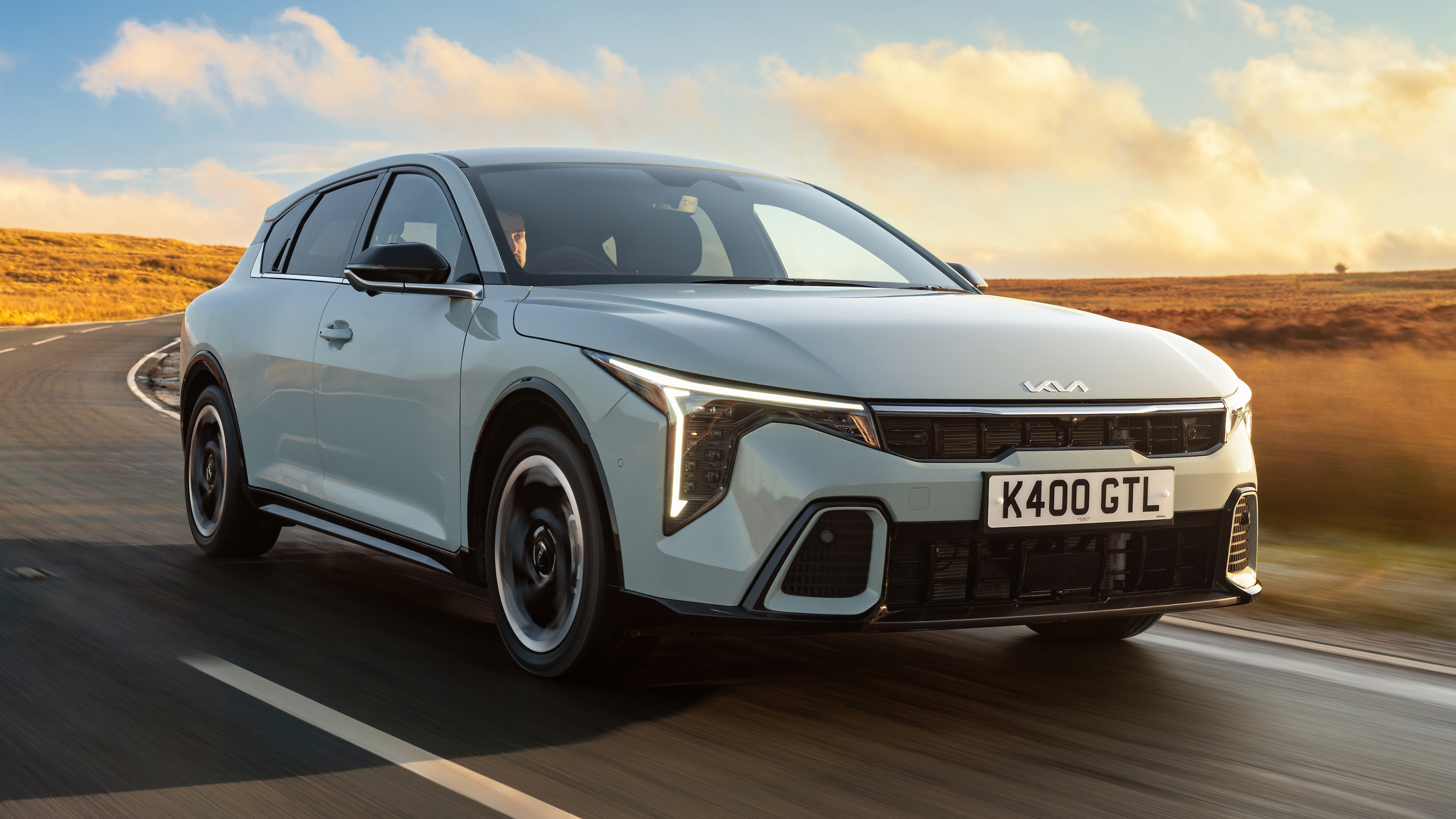Gray 2026 Kia K4 Hatchback driving on a countryside road at sunset.