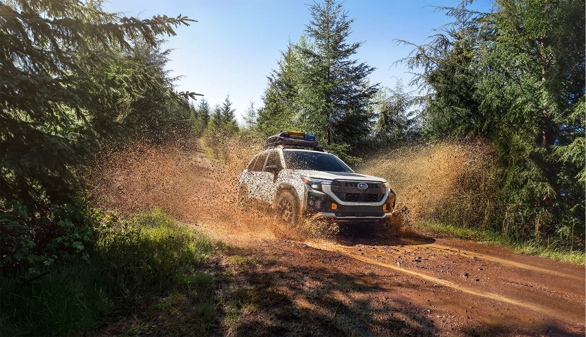 A white Subaru SUV driving through a muddy forest trail, splashing water and mud aggressively.