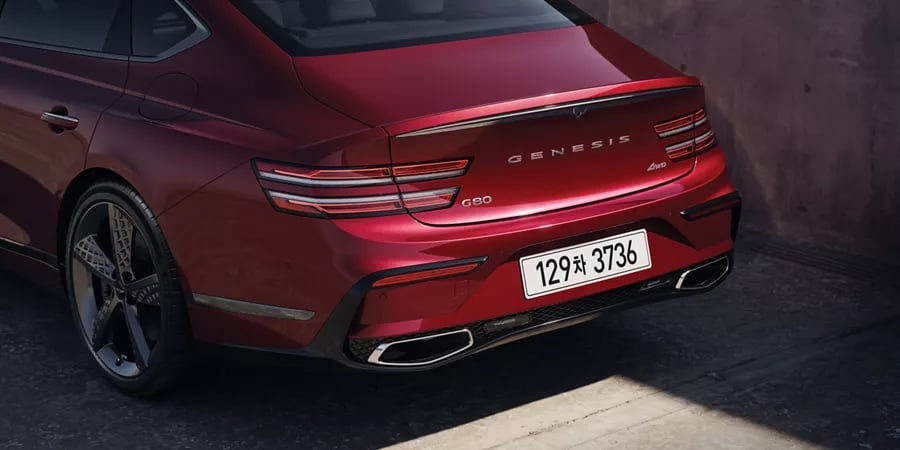 Rear view of a metallic red Genesis G80 luxury sedan showing signature LED tail lights and dual exhaust tips.
