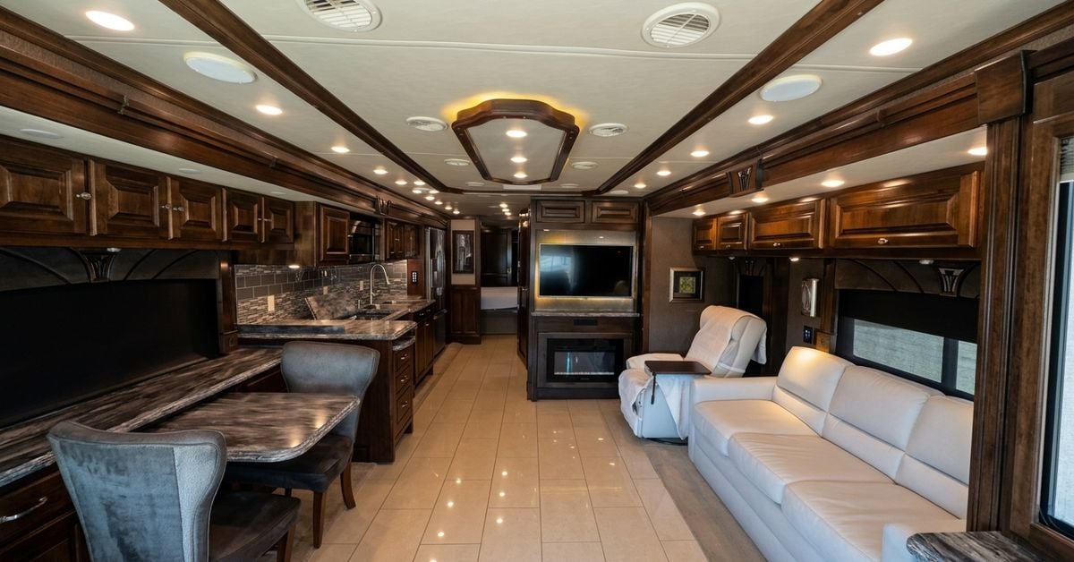 Spacious interior of a luxury motorhome featuring dark wood cabinetry, tile floors, a sofa, and a kitchen area.