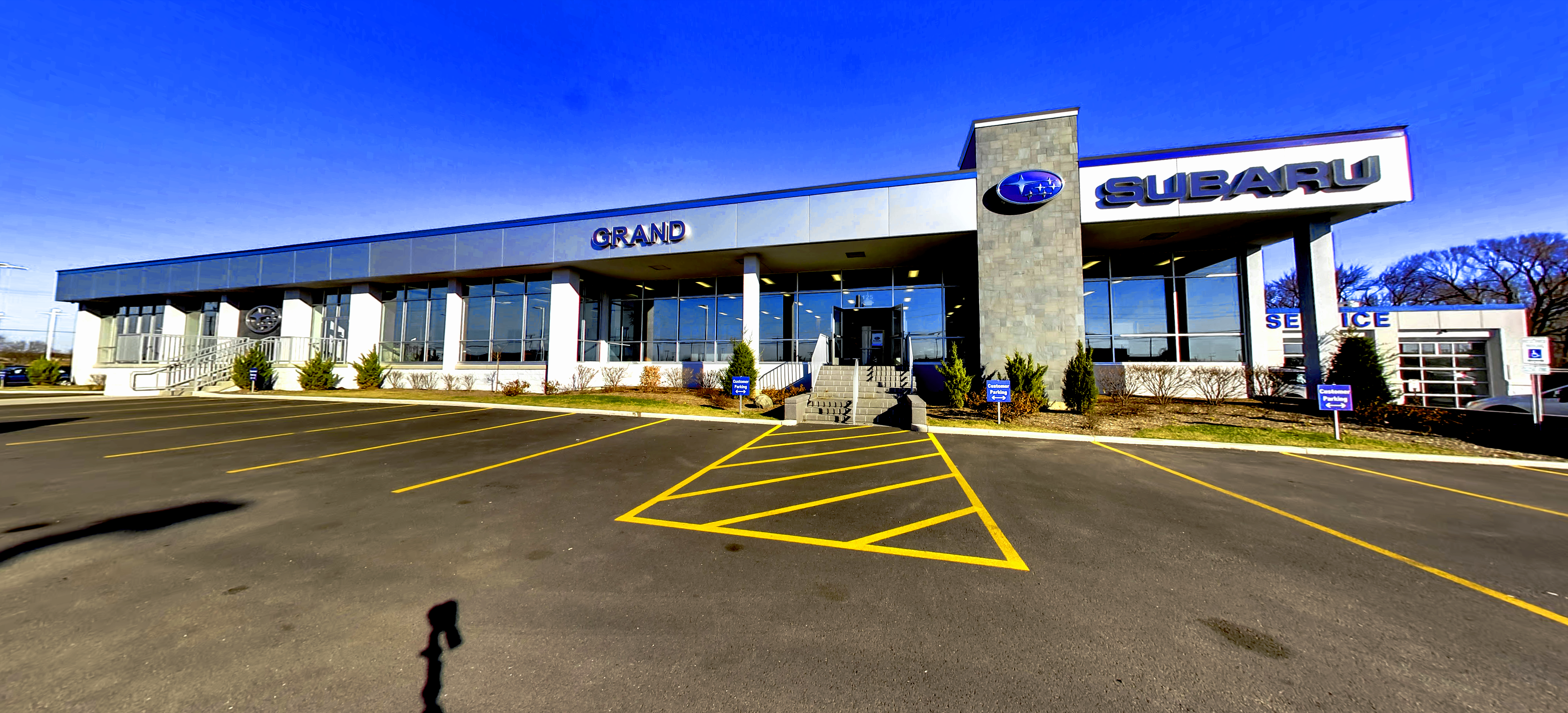 Wide shot of Grand Subaru dealership building and parking lot under a bright blue sky.