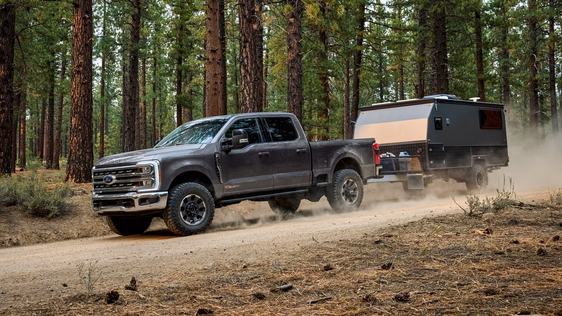 Gray Ford Super Duty truck towing camper on dirt road in forest.