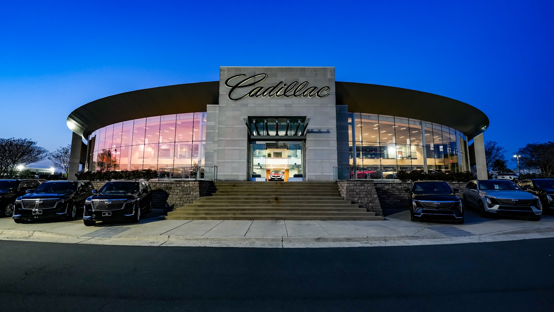 Exterior of a modern Cadillac dealership at dusk with large glass windows and luxury SUVs parked in front.