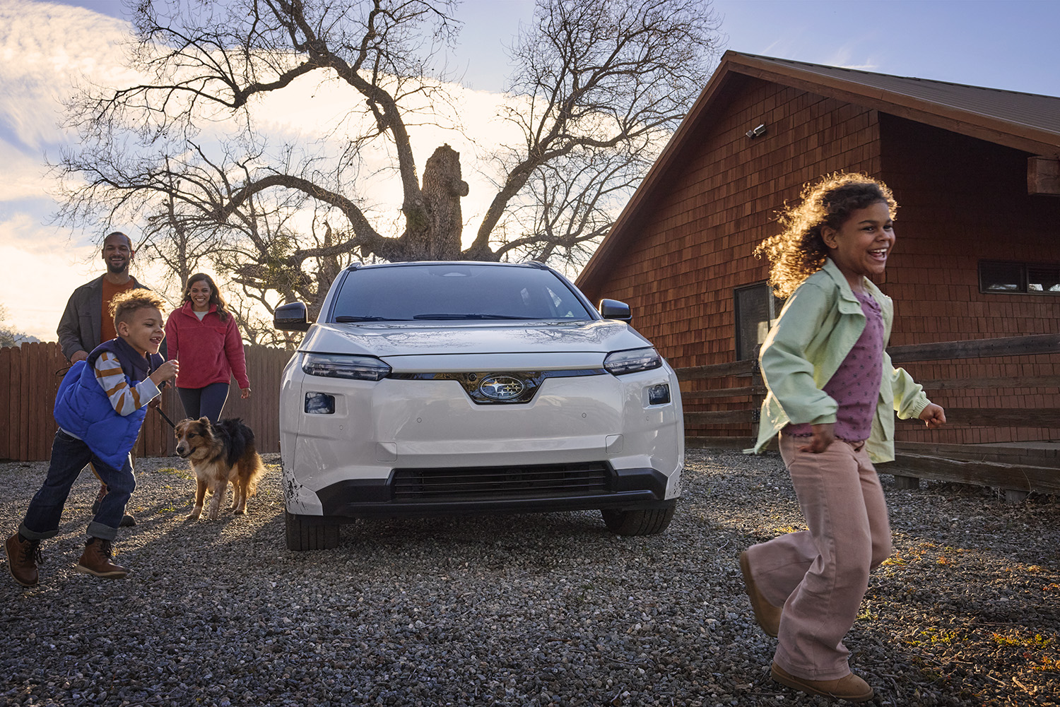 A happy family and their dog playing outdoors near a white Subaru Solterra electric SUV and a wooden cabin.
