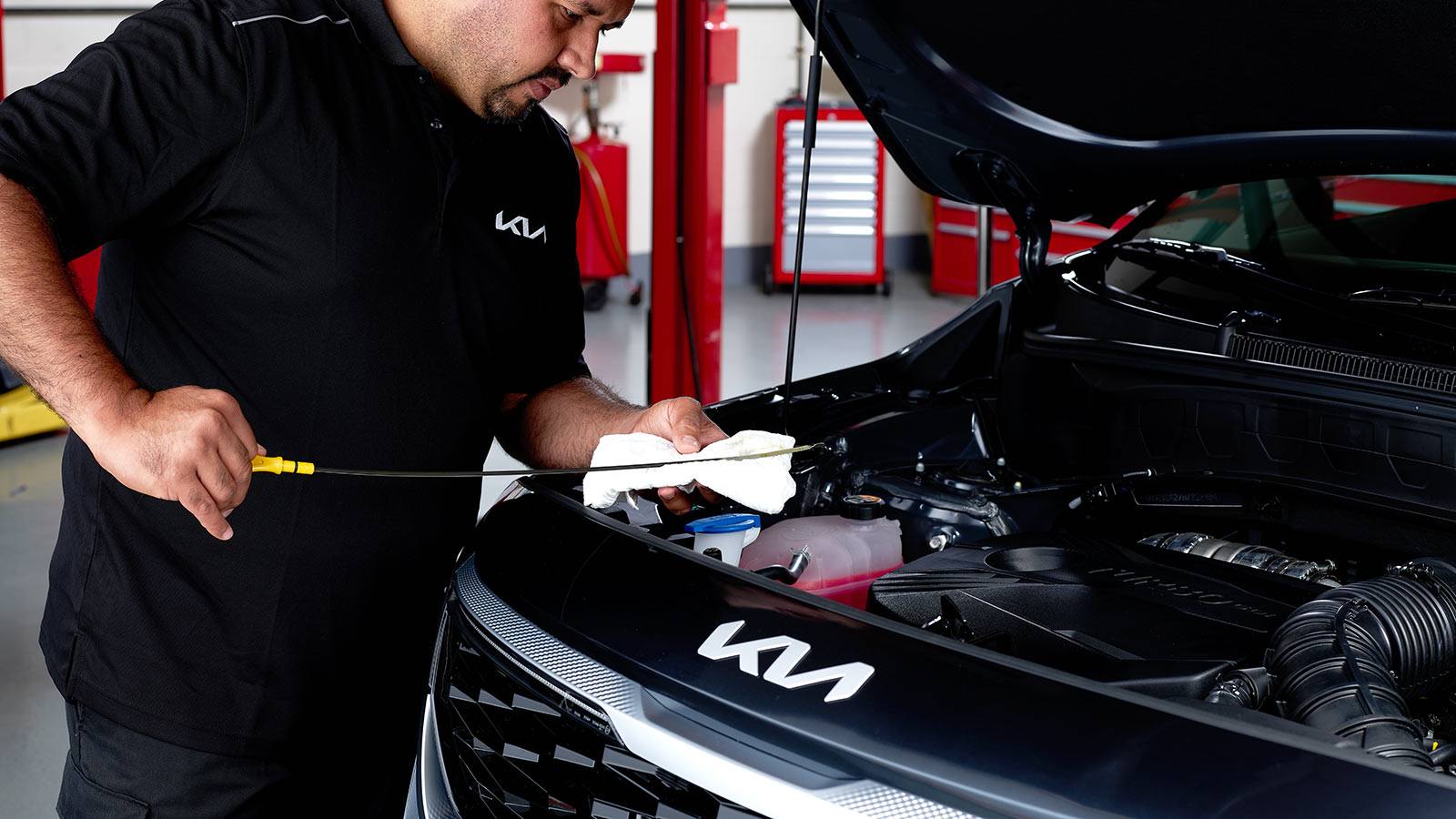 A Kia service technician pulls an engine oil dipstick and wipes it with a white cloth during car maintenance.