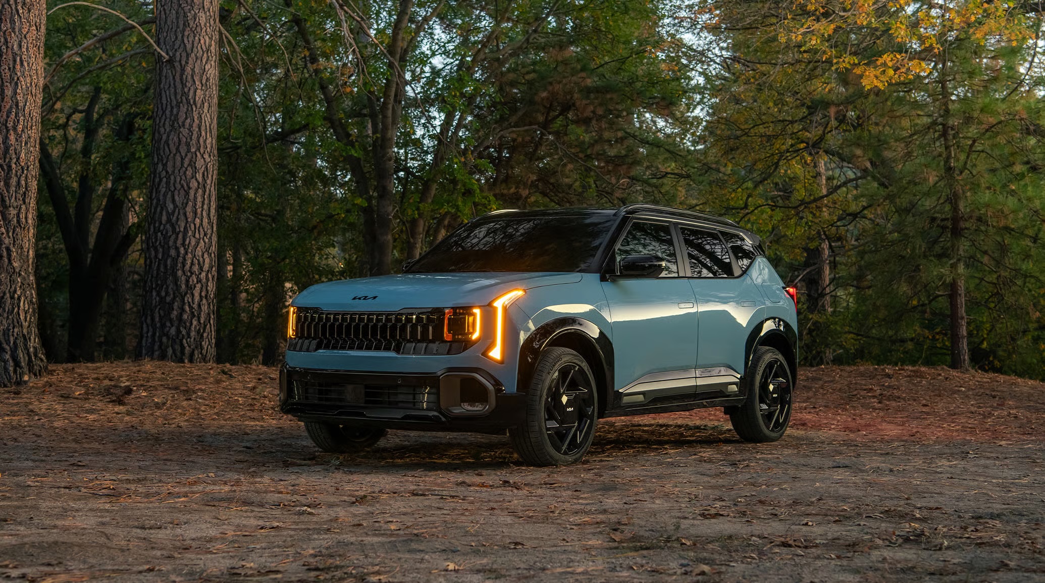 Front three-quarter view of a light blue Kia SUV with glowing amber headlights parked in a forest.