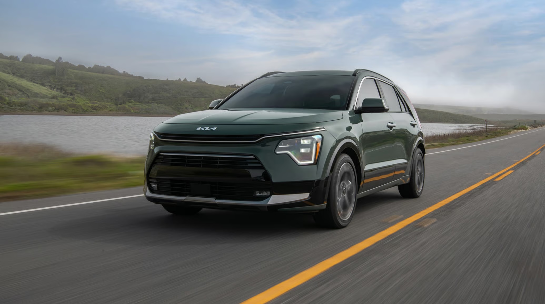 A dark green Kia Niro crossover SUV driving along a scenic two-lane coastal road.