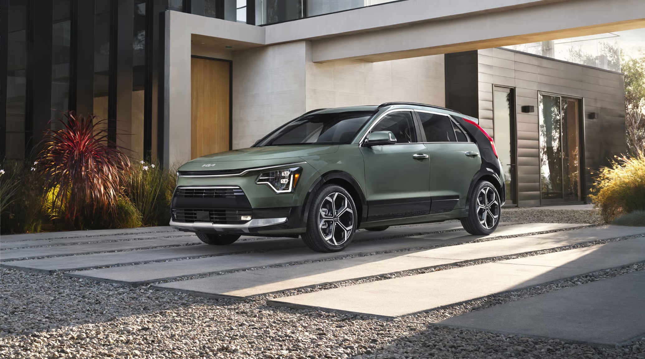 Sage green Kia Niro crossover parked on a concrete paver driveway outside a modern architectural home.