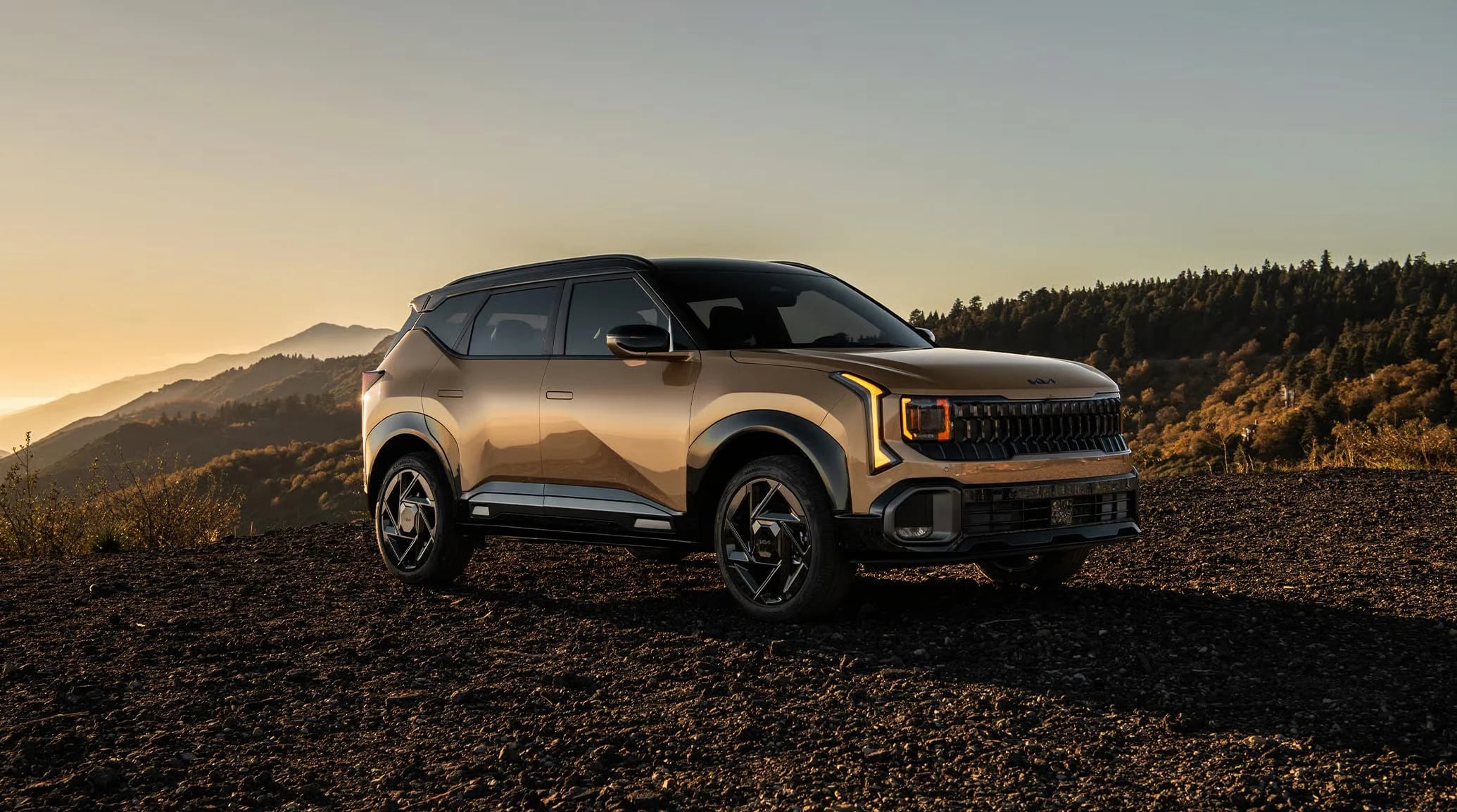 Metallic gold Kia compact crossover SUV parked on a rocky hilltop at sunset.