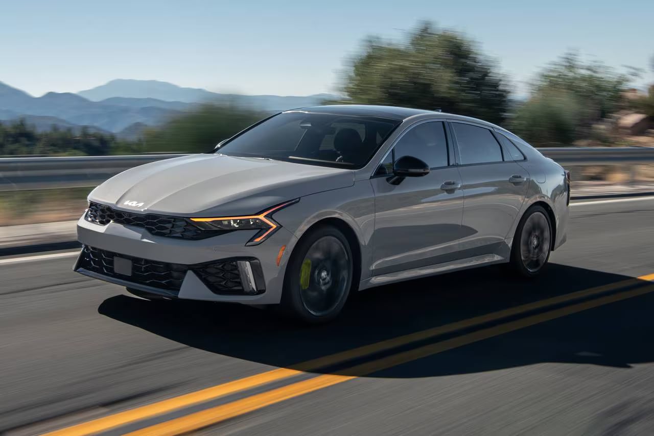 A light grey Kia K5 sedan driving fast along a paved road with amber LED lights glowing.