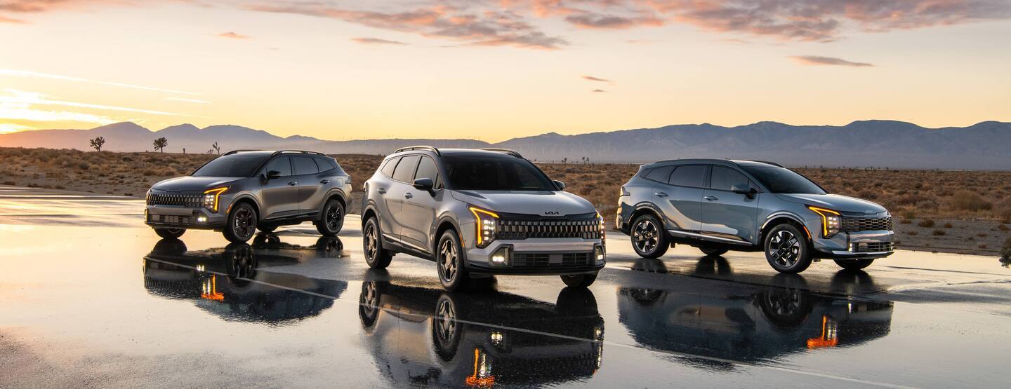 Three silver Kia Sportage SUVs parked in a line on wet pavement during sunset with a mountain backdrop.