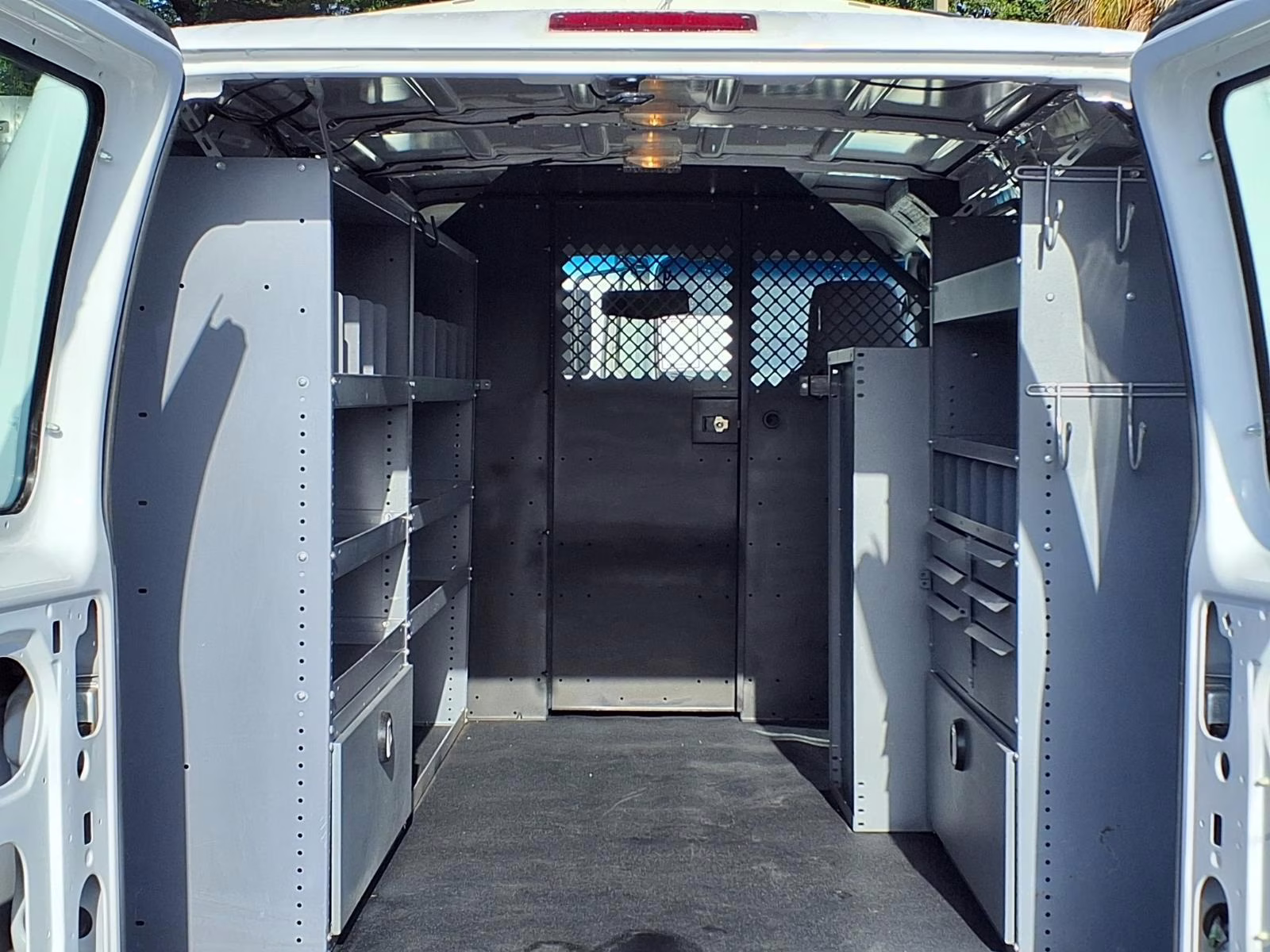 A professional white cargo van with interior shelving and lighting visible through the open side door, parked at a job site.