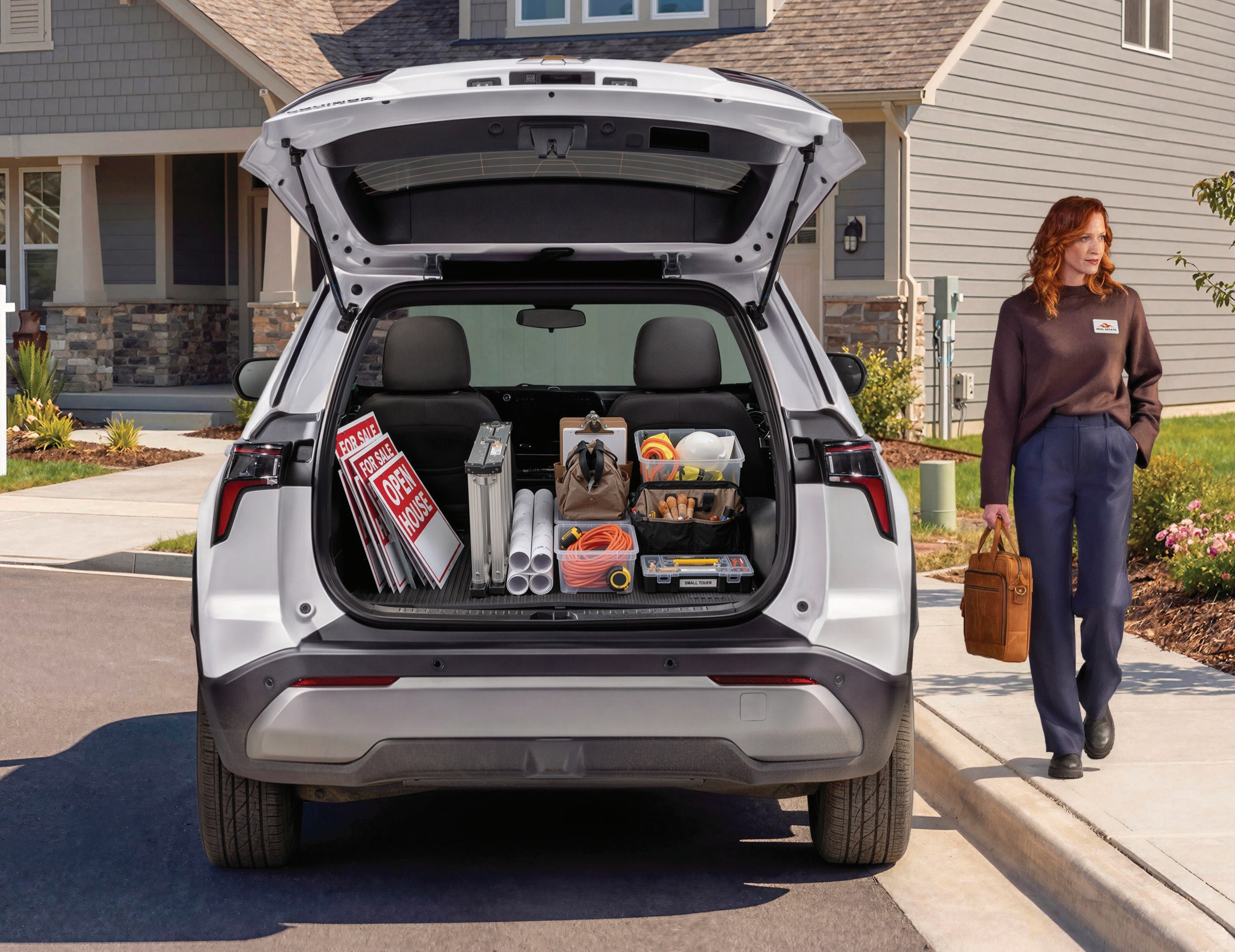 A woman walks past an open white SUV trunk filled with real estate signs, tools, and house supplies.