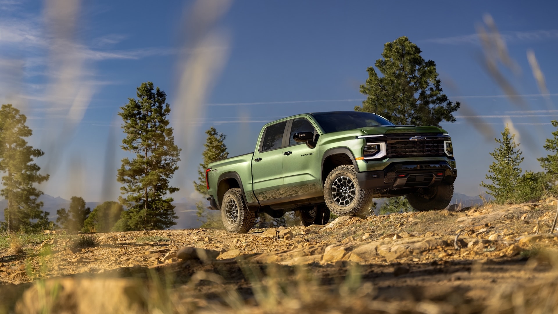 Metallic green Chevrolet Silverado EV off-road truck parked on a rocky dirt trail near pine trees.
