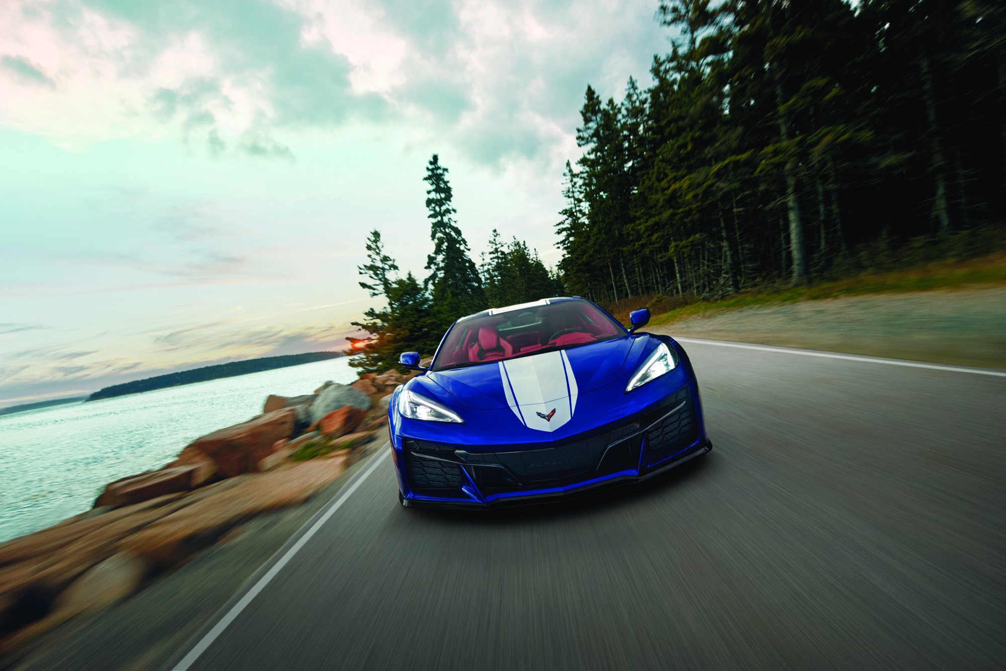 Blue Chevrolet Corvette with white racing stripes driving along a scenic coastal road at sunset.