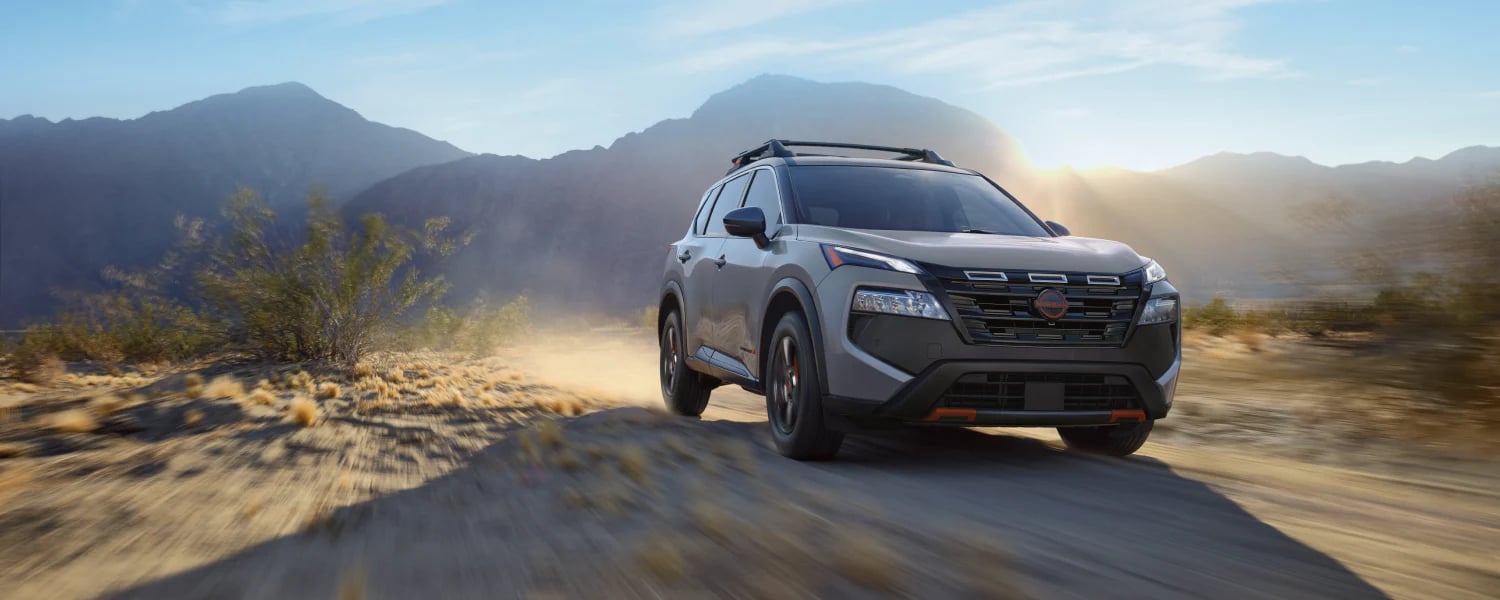 A silver Nissan SUV driving down a dusty desert trail with sunlit mountains in the background.