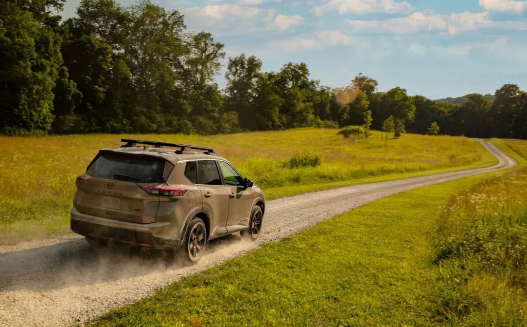 A bronze Nissan Rogue SUV driving down a gravel road through a grassy field, kicking up dust in sunny weather.